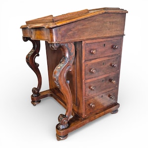 May include: An antique wooden writing desk with a sloping top and drawers. The desk has ornate, carved legs and a dark brown finish. The drawers have small, round knobs. The desk is likely made of mahogany.