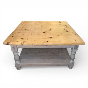 May include: Square wooden coffee table with a light brown top and a grey base. The table has a lower shelf and decorative turned legs. The top is approximately 91 cm square.