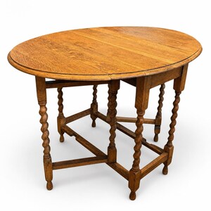 May include: An oval drop-leaf table crafted from medium-toned oak wood. The table features a smooth, flat tabletop surface with visible wood grain patterns and is supported by six legs, each designed with a distinct twisted, spiral-turned column detail.