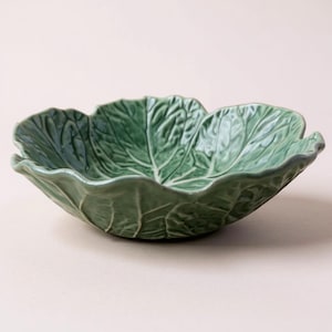 May include: A green ceramic serving bowl in the shape of a cabbage leaf. The bowl features detailed veining and a slightly ruffled edge. The bowl is a solid green colour and is set against a neutral background.
