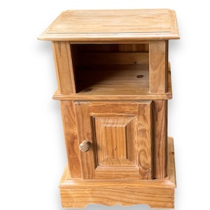 May include: A wooden bedside table with a cabinet door and an open shelf. The table is made of light-coloured wood and has a simple, rustic design. The cabinet door has a small, round wooden knob.