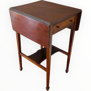 May include: A dark brown, antique wooden side table with a drop-leaf design. The table features a single drawer with a black knob and a lower shelf. The legs are slender and tapered, with small decorative feet. The table is approximately 71 cm tall.