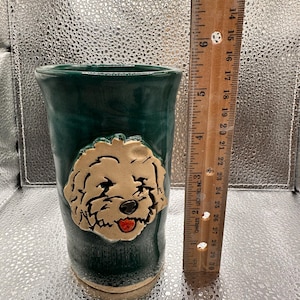 May include: A handcrafted, dark teal ceramic tumbler with a sculpted, tan-colored dog face. The dog has black details and a red tongue. The tumbler is approximately 6 inches tall.
