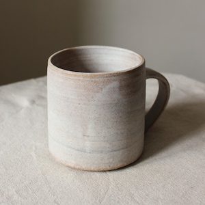 Könnte beinhalten: Eine handgefertigte, cremefarbene Keramik-Tasse mit rundem Henkel. Die Tasse hat eine zylindrische Form und eine leicht strukturierte Oberfläche. Der Innenraum ist in einem helleren Beigeton gehalten. Die Tasse eignet sich für heiße oder kalte Getränke.