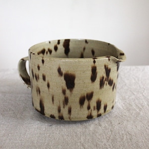 Può includere: Brocca in ceramica beige con macchie marrone scuro, manico e beccuccio. La brocca sembra fatta a mano. Il manico è curvo e il beccuccio leggermente angolato. Adatta per servire liquidi, probabilmente un articolo per la cucina.