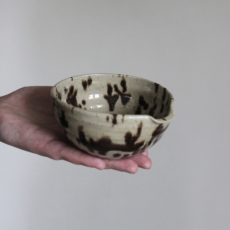 A small, brown speckled ceramic bowl with a pouring spout. The bowl has a speckled glaze with brown splotches and streaks.
