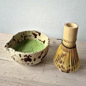 Speckled stoneware matcha bowl paired with bamboo whisk and mat ha powder, photographed in natural light.