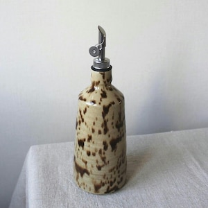 May include: A ceramic oil bottle with a stainless steel spout. The bottle is beige with brown speckled patterns. The spout is silver and has a curved design. The bottle is designed for dispensing oil or vinegar.