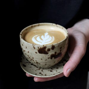 Op de afbeelding: Een keramieken kop en schotel met een gespikkelde beige afwerking en donkerbruine vlekken. De kop is gevuld met een latte met een wit, bladvormig schuimontwerp. Het serviesstuk heeft een afgeronde vorm en een textuurrijk, aards uiterlijk.