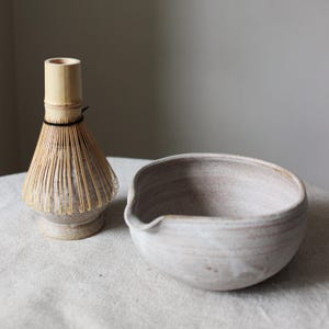Op de afbeelding: Een witte keramische kom met een tuit en een bamboe garde met een bruine keramische voet. De kom en de garde staan op een wit linnen oppervlak.