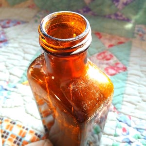 May include: A rectangular amber glass bottle with a narrow neck and a threaded top. The glass is translucent and brown, featuring embossed lettering on the side that is partially visible. The bottle has a distinct shoulder and a flat base.