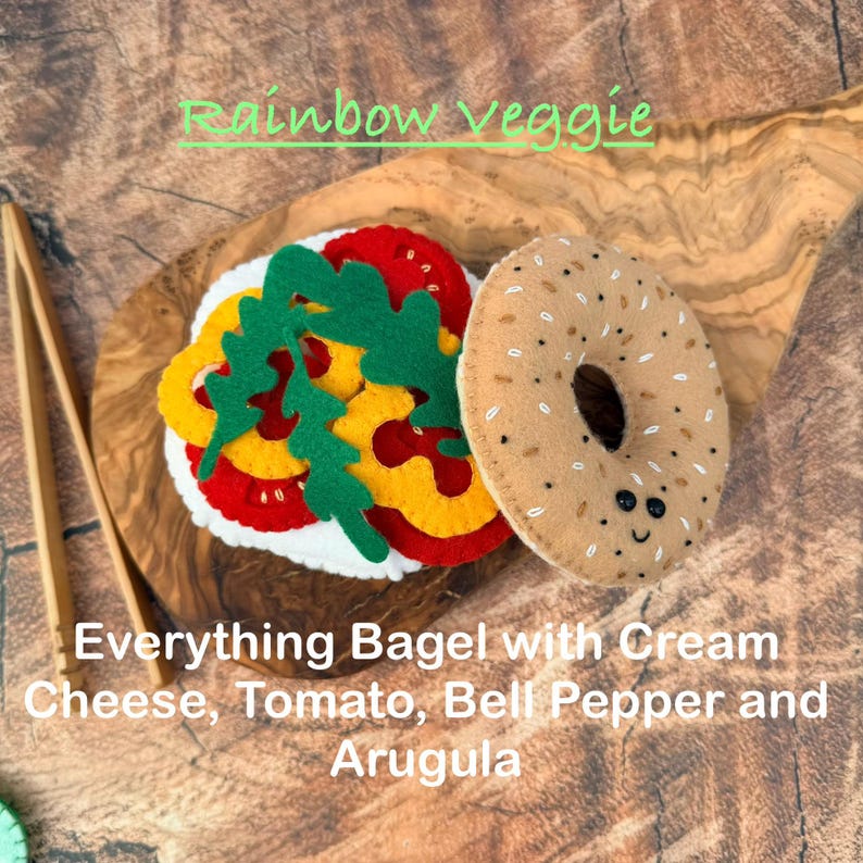 May include: A felt play food set featuring an everything bagel with a smiling face, layered with white cream cheese, red tomato slices, yellow bell pepper rings, and green arugula leaves. The text Rainbow veggie and Everything Bagel with Cream Cheese, Tomato, Bell Pepper and Arugula is visible.