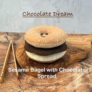 May include: A felt plush toy shaped like a bagel sandwich with a tan top featuring white embroidered sesame seeds and a small smiling face, a dark brown middle layer, and a cream bottom layer, labeled Chocolate Dream and Sesame Bagel.