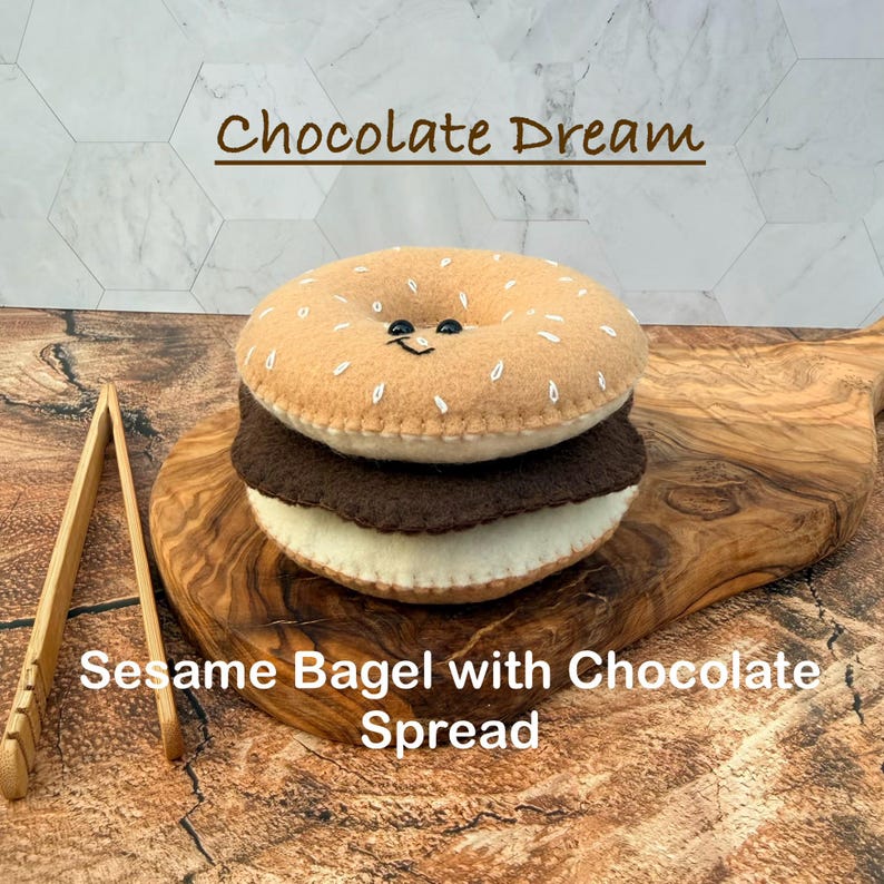 May include: A felt plush toy shaped like a bagel sandwich with a tan top featuring white embroidered sesame seeds and a small smiling face, a dark brown middle layer, and a cream bottom layer, labeled Chocolate Dream and Sesame Bagel.