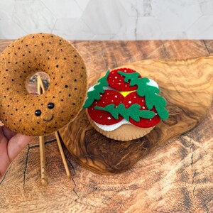 May include: A tan felt bagel with black speckles and a stitched smiling face, positioned next to a felt food item featuring a white base, red circular toppings, and green leaf-shaped accents, all crafted from soft textile materials.