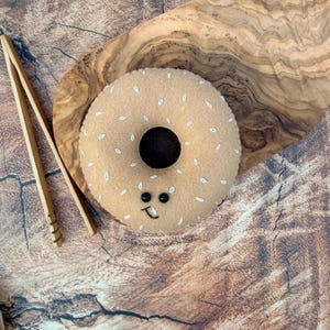 May include: A tan felt donut toy featuring white embroidered seed details, two small black bead eyes, and a stitched black mouth. The donut is positioned next to a pair of light brown wooden kitchen tongs on a textured wooden surface.