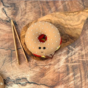 May include: A felt bagel toy with a tan exterior, white seed-like accents, and two small black bead eyes with a stitched smile. The bagel is layered with red, yellow, and brown felt fillings, accompanied by a pair of light-colored wooden tongs.