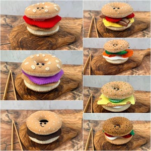 May include: A collection of felt bagel sandwiches featuring various fillings including red, purple, yellow, and brown layers. Each bagel has a tan top with embroidered white seeds and a stitched smiling face with black eyes and a curved mouth.