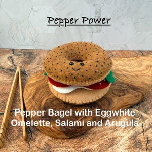 May include: A felt toy shaped like a bagel with a smiling face, featuring black speckles, a white egg white omelette, red salami, and green arugula. The text Pepper Power and Pepper Bagel with Eggwhite Omelette, Salami and Arugula is visible.