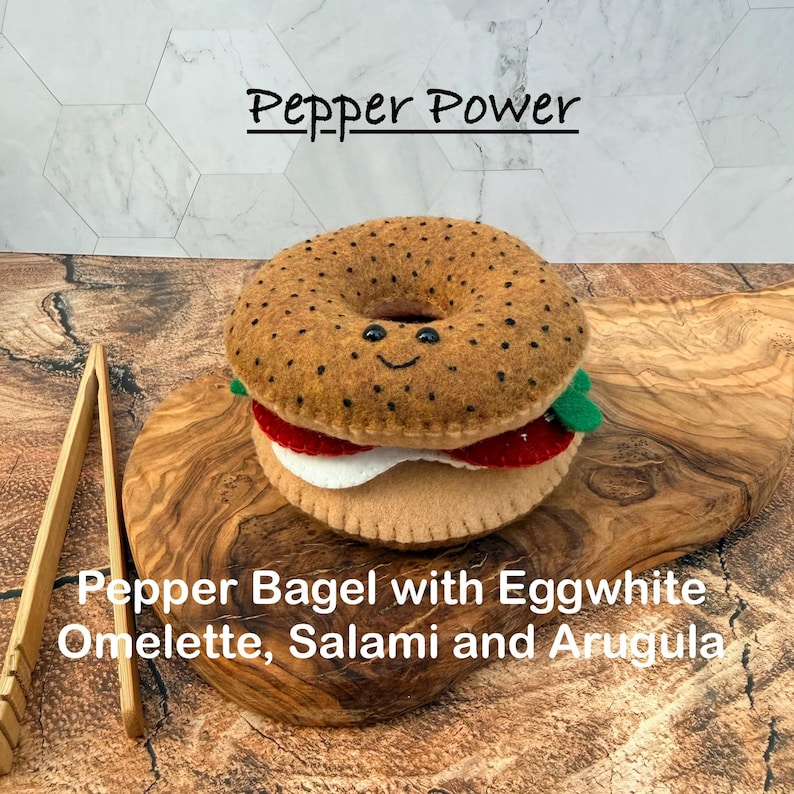 May include: A felt toy shaped like a bagel with a smiling face, featuring black speckles, a white egg white omelette, red salami, and green arugula. The text Pepper Power and Pepper Bagel with Eggwhite Omelette, Salami and Arugula is visible.