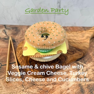 May include: A felt bagel toy with embroidered green sesame seeds and a smiling face, layered with yellow cheese, green cucumber, and white cream cheese. The text Garden Party and Sesame & chive Bagel with Veggie Cream Cheese, Turkey Slices, Cheese and Cucumbers is visible.