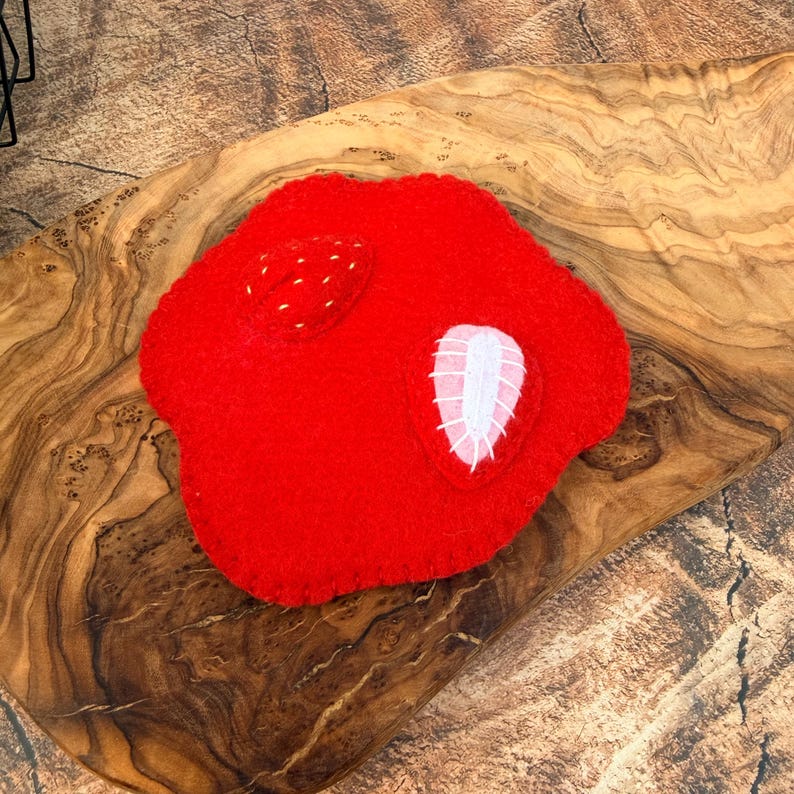 May include: A bright red felted textile piece shaped like a rounded, irregular form. It features two small, stitched strawberry-like appliqu&eacute;s on its surface: one is a solid red textured shape, and the other is a white oval with red stitching.
