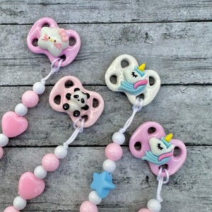 以下が含まれることがあります： 装飾的なチャームが付いた4つのベビーおしゃぶり。2つのピンクのおしゃぶりには、ハローキティとパンダのチャームが付いています。他の2つのおしゃぶりは、ユニコーンのチャームが付いた白いベースです。各おしゃぶりはビーズチェーンに接続されています。