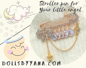 Può includere: Spilla dorata per passeggino con un fiocco, ciondoli e il nome "ANGEL" scritto. La spilla presenta anche una croce e un ciondolo con l'occhio turco. Il testo "DOLLSBYYANA.COM" è visibile nella parte inferiore dell'immagine.