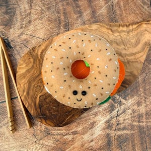 May include: A tan felt bagel toy with embroidered brown and white seed details, featuring two small black bead eyes and a stitched smiling mouth. The bagel has a visible orange and green felt filling layer and is paired with wooden tongs.