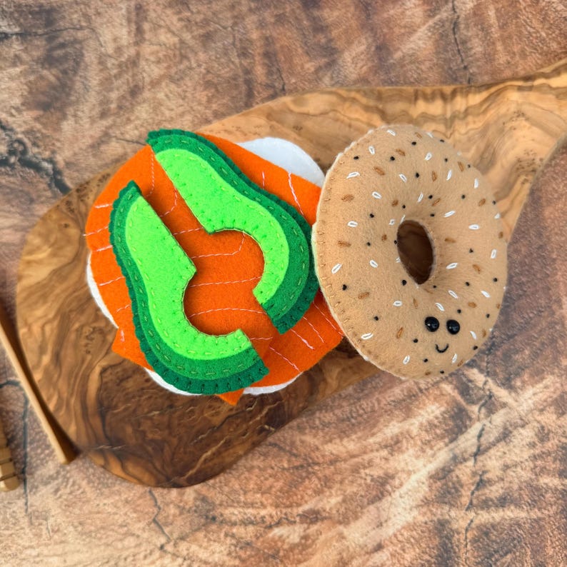 May include: A felt toy set featuring a tan bagel with embroidered seeds and a small smiling face, paired with a stack of white bread, orange salmon slices, and bright green avocado wedges with dark green edges, all crafted from soft felt fabric.