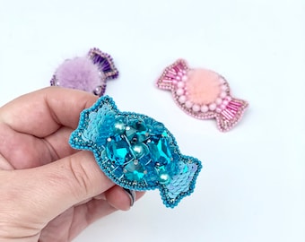Puede incluir: Un broche azul en forma de caramelo con lentejuelas y cuentas. El broche está hecho de lentejuelas y cuentas azules, y tiene un pompón blanco en el centro.