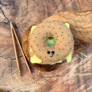 May include: A tan felt bagel toy with embroidered green and white details, featuring a smiling face with black bead eyes and a stitched mouth. The bagel is layered over a yellow felt square and includes a pair of light brown wooden tongs.
