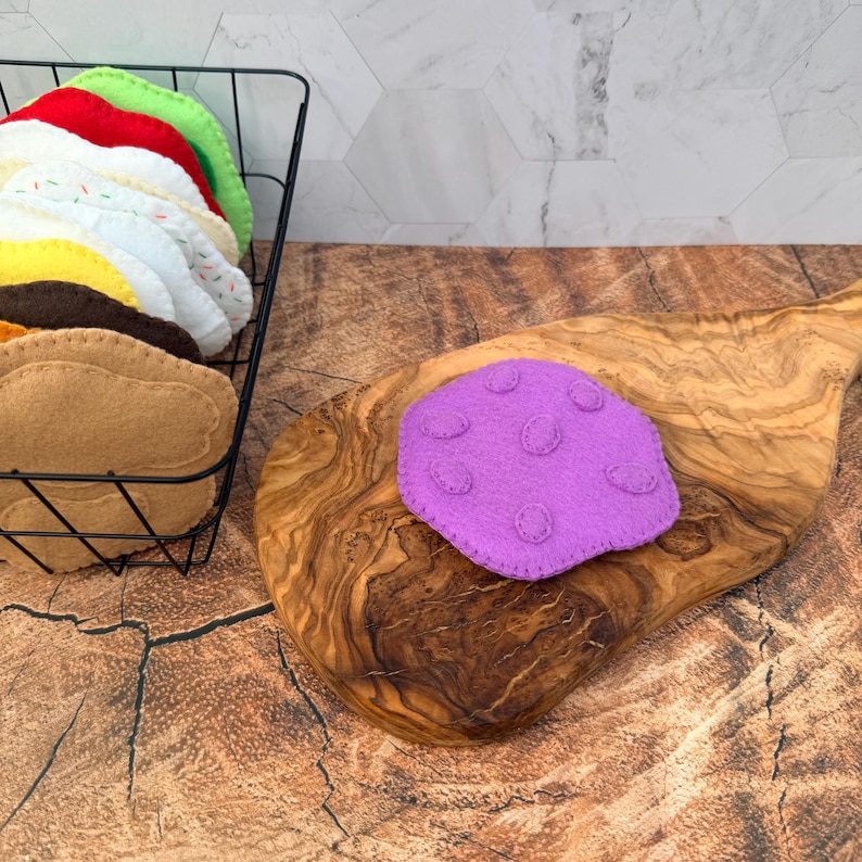 May include: A purple felt cookie with small circular felt accents sits on a wooden cutting board. A black wire basket holds a stack of various colored felt cookies, including red, green, white, yellow, and brown, with visible stitching edges.