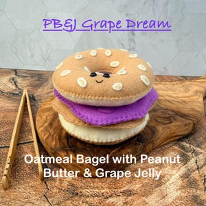 May include: A felt plush toy shaped like a bagel sandwich with peanut butter and grape jelly. The top bagel half is tan with white accents and a smiling face. The middle layer is purple, and the bottom layer is tan. Text reads PB&J Grape Dream.