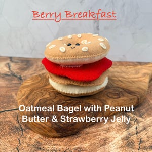 May include: A felt toy shaped like a bagel sandwich with a tan top featuring embroidered oats and a smiling face, a red jelly layer, and a tan bottom. The text Berry Breakfast and Oatmeal Bagel with Peanut Butter & Strawberry Jelly is visible.
