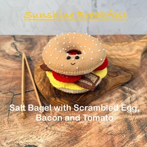 May include: A felt bagel sandwich with a smiling face, topped with white salt crystals. The sandwich includes layers of yellow scrambled egg, red tomato, and brown bacon. A pair of wooden tongs sits beside the bagel. Text reads Sunshine Breakfast.