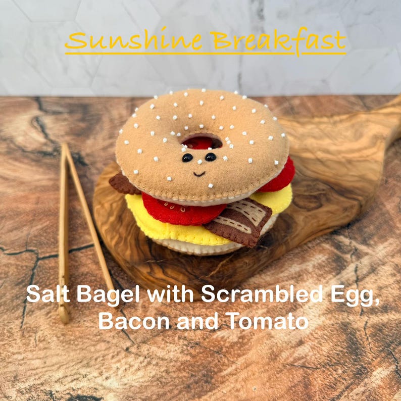 May include: A felt bagel sandwich with a smiling face, topped with white salt crystals. The sandwich includes layers of yellow scrambled egg, red tomato, and brown bacon. A pair of wooden tongs sits beside the bagel. Text reads Sunshine Breakfast.
