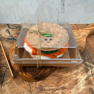May include: A felt bagel sandwich toy with a smiling face, featuring tan, orange, and white layers, tied with brown twine inside a clear plastic container. The bagel has embroidered details and is presented as a single decorative item.