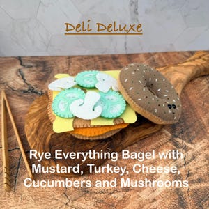 May include: A felt toy rye everything bagel sandwich featuring layers of yellow cheese, orange turkey, light green cucumber slices, and white mushroom pieces. The text Deli Deluxe and Rye Everything Bagel with Mustard, Turkey, Cheese, Cucumbers and Mushrooms is visible.