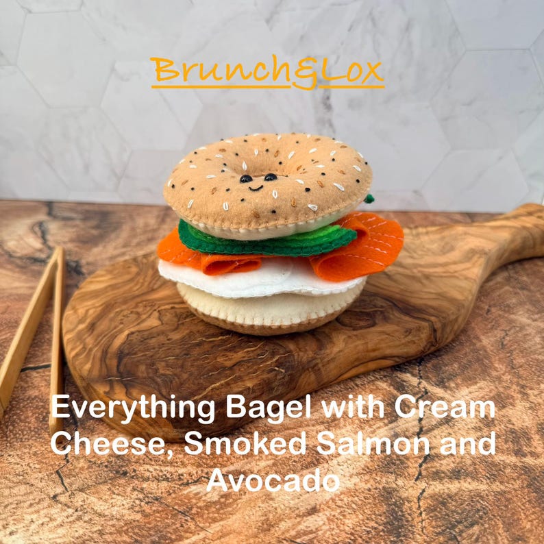 May include: A felt bagel sandwich featuring a tan top bun with black and white speckles, a green avocado slice, orange smoked salmon, and a white cream cheese layer. The text reads Brunch&Lox and Everything Bagel with Cream Cheese, Smoked Salmon and Avocado.