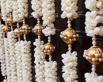 Puede incluir: Una guirnalda decorativa colgante con múltiples hilos verticales de flores de jazmín artificiales blancas. Cada hilo está acentuado con cuentas metálicas doradas y pequeñas perlas blancas, creando una exhibición floral texturizada.