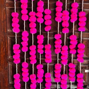 Puede incluir: Guirnaldas de pompones rosa brillante colgadas frente a una puerta de madera marrón oscuro. Cada guirnalda está hecha de múltiples pompones ensartados con una cuerda blanca. La puerta tiene un diseño tradicional con manija y cerradura.