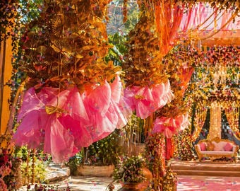 May include: An outdoor wedding or event setting with elaborate decorations. Gold leaf and pink fabric hang from the ceiling. Floral arrangements and lanterns line the pathway. A sofa is visible in the background, creating a festive atmosphere.