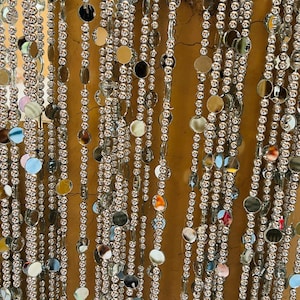 Può includere: Tenda decorativa di perline con fili di piccole perline argentate lucenti e dischi circolari riflettenti più grandi. La tenda è appesa in file verticali, creando un effetto scintillante. Lo sfondo è di un colore caldo e neutro.