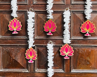 May include: Door decorations with pink lotus flower designs and gold accents, suspended from a dark brown wooden door. White garland is used to hang the lotus flowers. The door is a dark brown colour.
