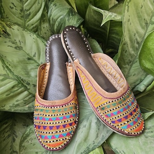 May include: A pair of colorful embroidered slip-on shoes. The shoes feature a brown leather sole and a beige upper with intricate stitching in shades of orange, blue, green, and pink. The shoes are set against a backdrop of green leaves.