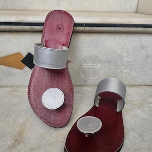May include: Burgundy leather sandals with silver detailing. Each sandal has a silver strap and a circular silver feature. The sandals are handmade.