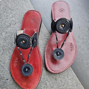 Peut inclure: Une paire de sandales plates avec des semelles en cuir rouge foncé et des brides noires. Les brides sont ornées de médaillons circulaires à bords festonnés et d'un rivet en laiton au centre. La taille 39 est imprimée sur le talon.