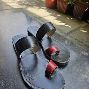 May include: A pair of black leather sandals featuring a wide strap across the midfoot and a separate red leather toe loop. The flat soles are stitched along the edges, and the brand name is embossed on the heel area of the footbed.