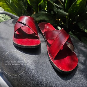 May include: A pair of handmade red leather sandals with a crisscross strap design. The sandals are displayed on a black textured surface, with green foliage in the background. The text "THIS PRODUCT IS 100% HANDMADE" is visible.